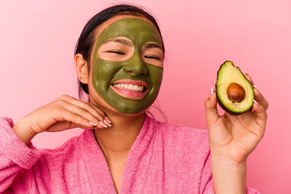 Mujer joven sonriente mostrando en su mano medio aguacate mientras se aplica en el rostro una mascarilla casera hidratante