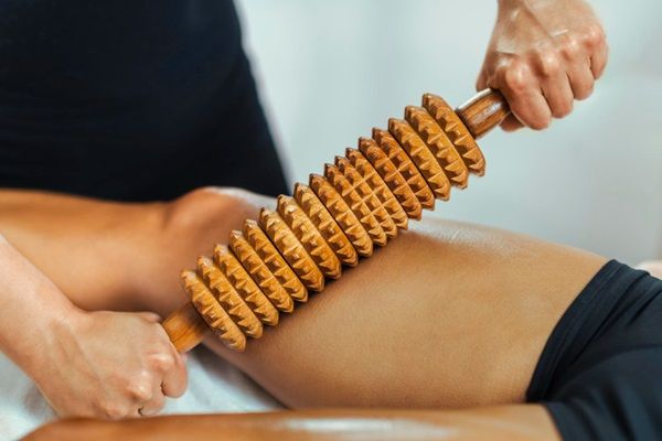 Médico estético realizando un masaje con un rodillo estriado de madera en las piernas de un paciente