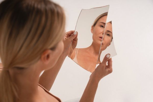 Tratamiento estético - Mujer mirándose en un espejo roto con baja autoestima por imperfecciones faciales que se pueden resolver con tratamientos estéticos